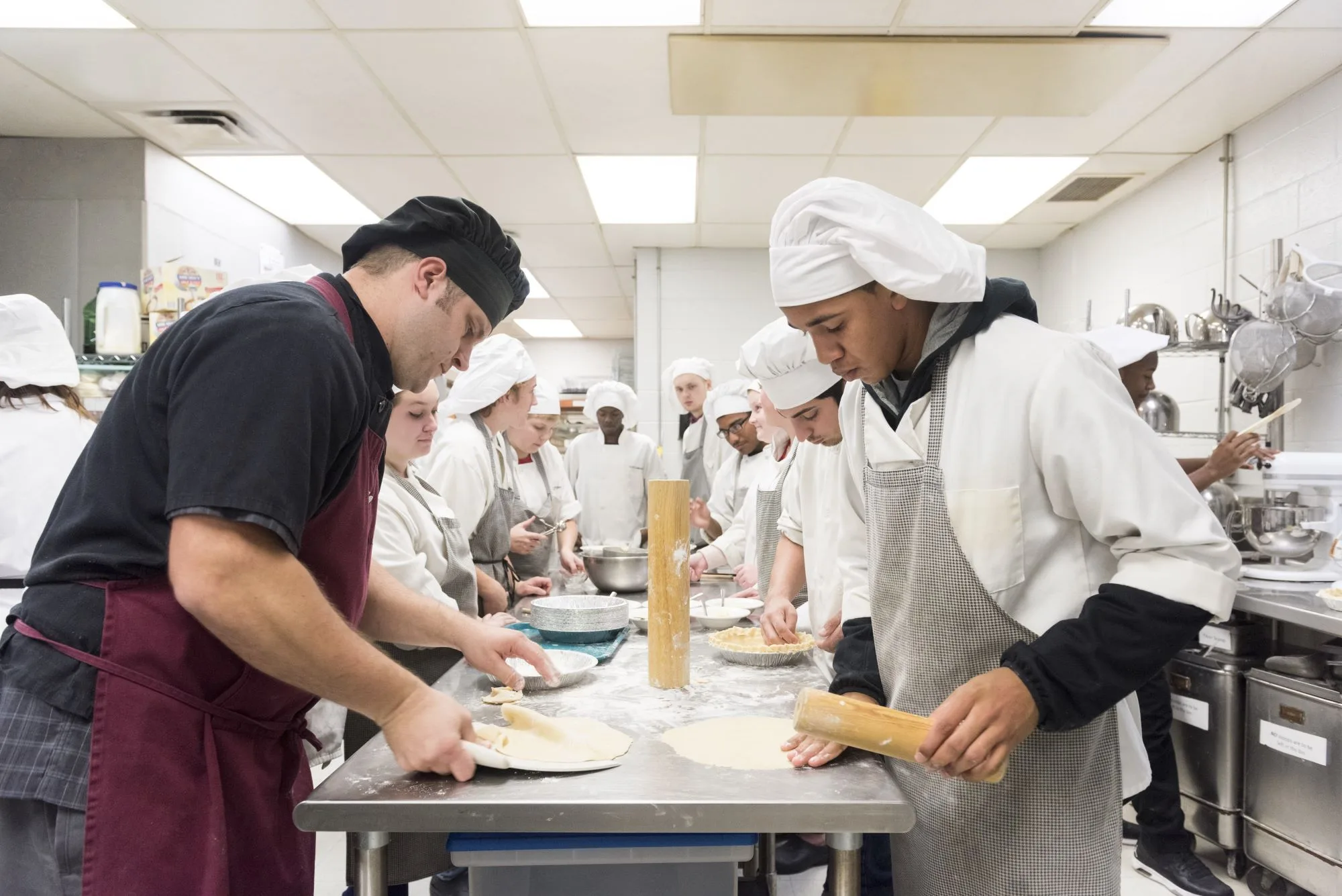 Chef instructor guides students rolling and shaping pie dough in a commercial kitchen.