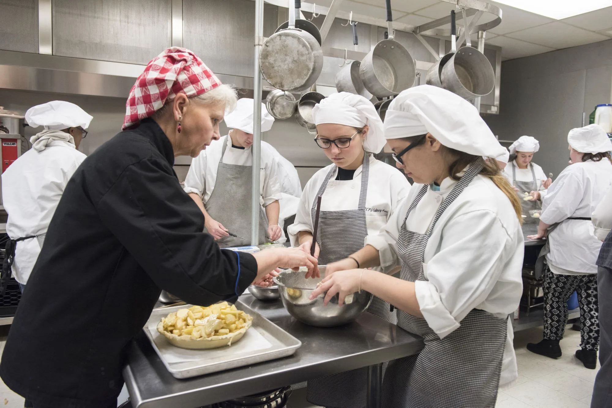 Chef in a red checkered bandana guides students with an apple pie in a kitchen.