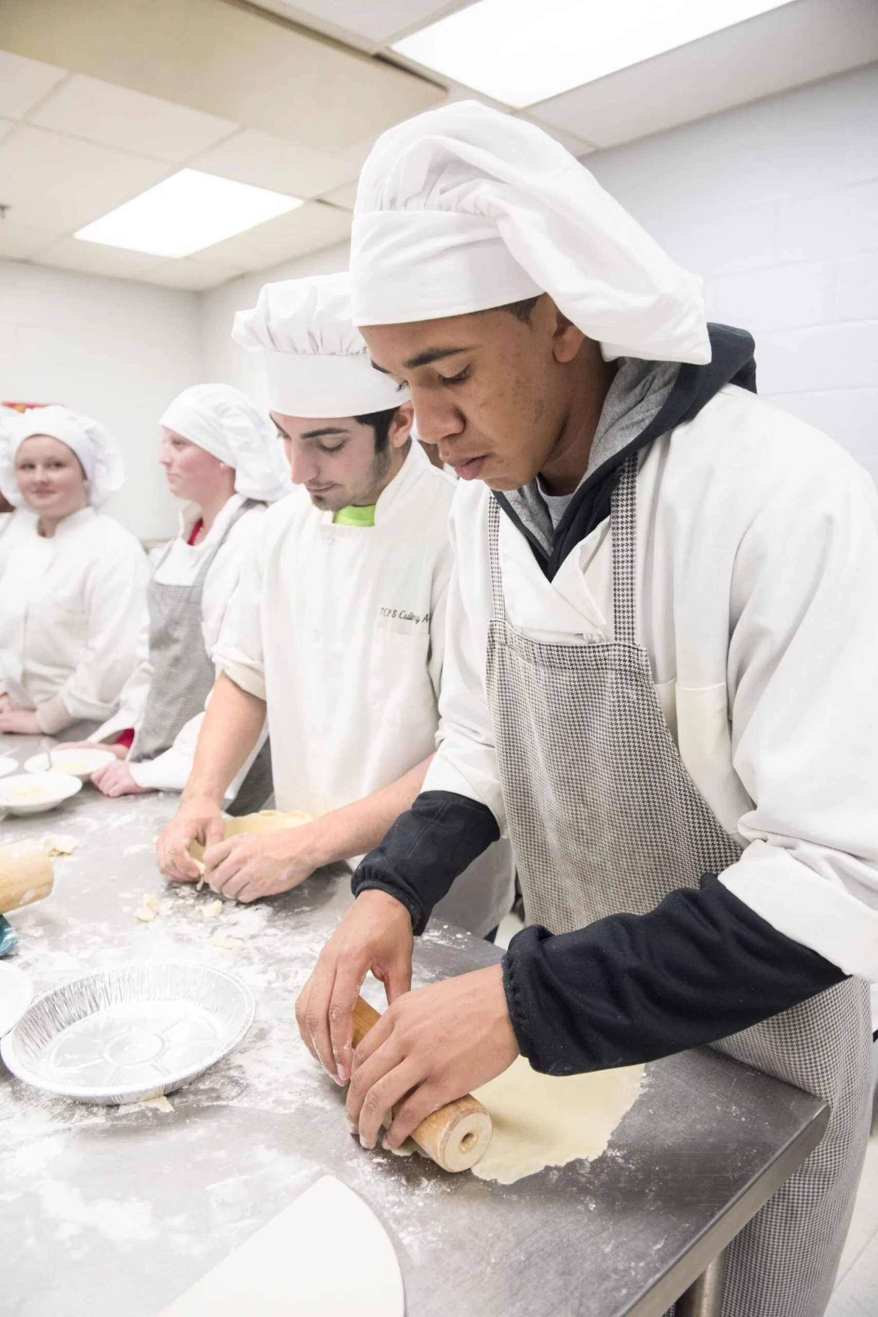 Student in chef whites rolls out pie dough on a floured counter.