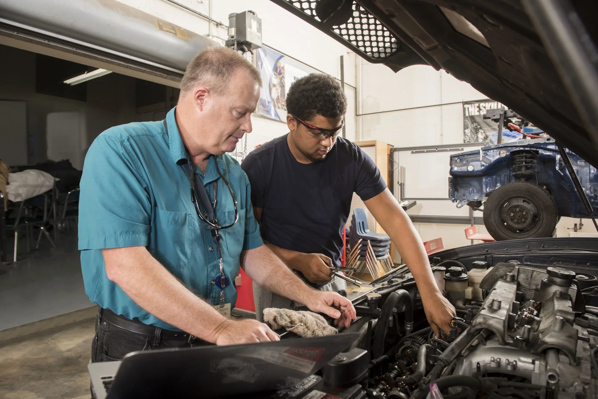 Instructor in teal shirt helps a student work under a car hood.
