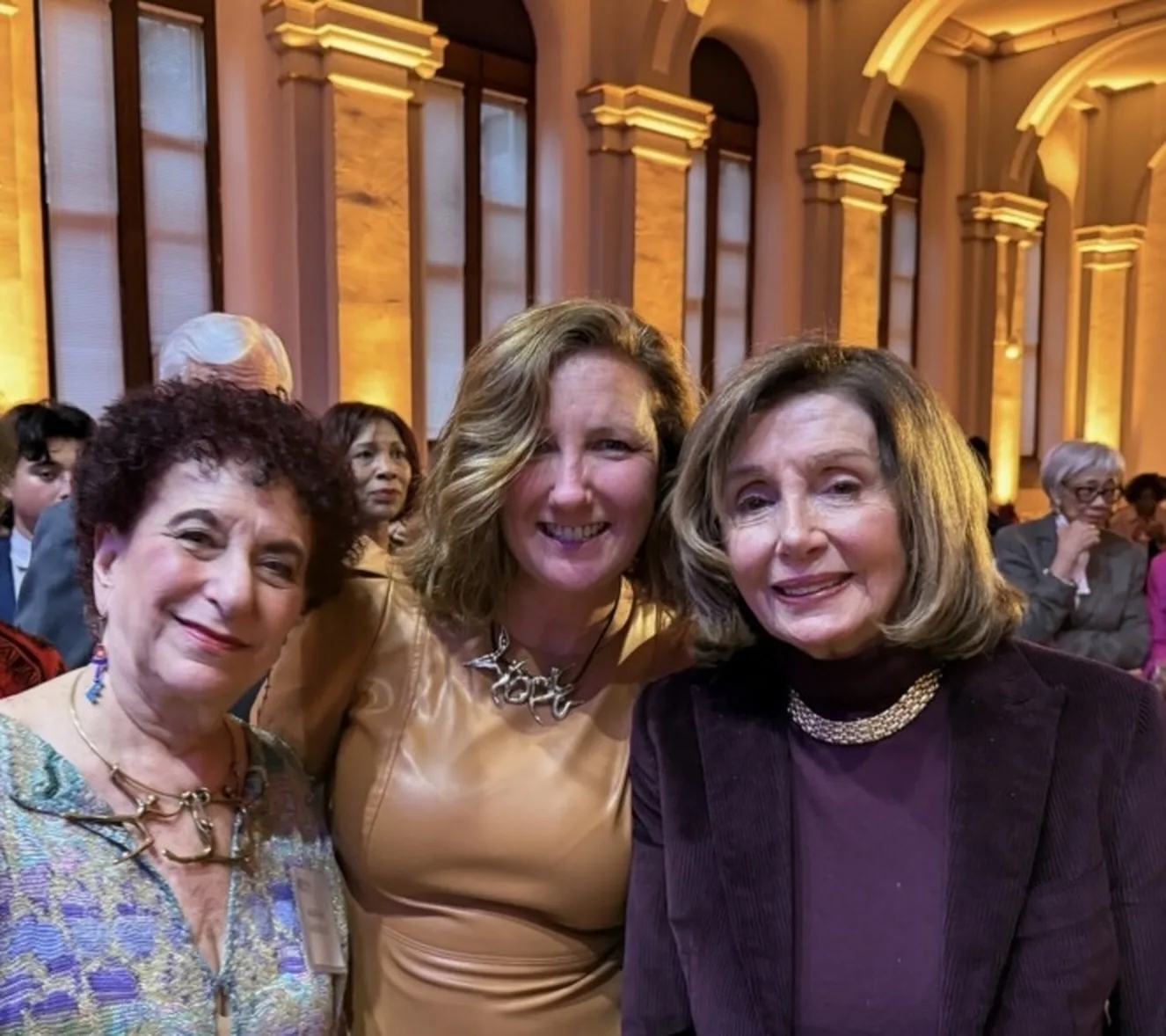 Three women smile together in an ornate, gold-lit hall.