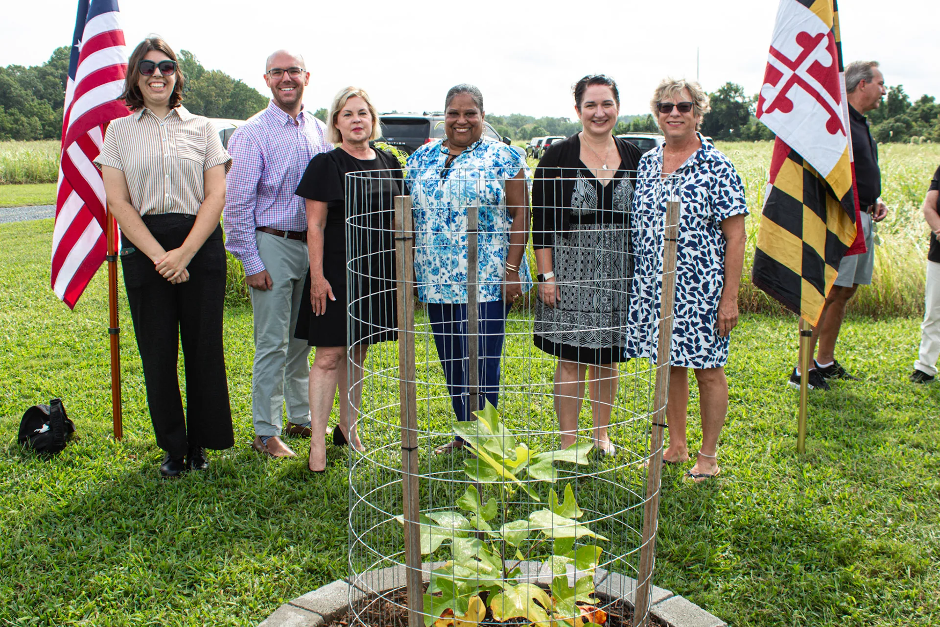 Six smiling people posing beside a wire-caged sapling, American and Maryland flags on either side
